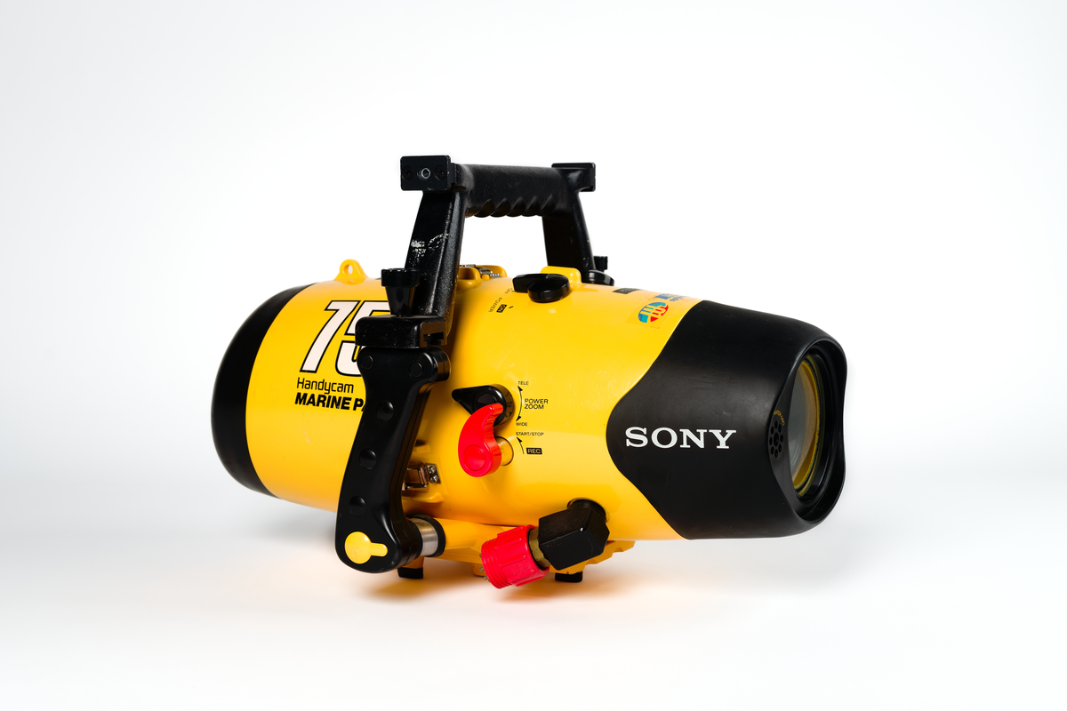 Videokamera i undervattenshus. Gult plasthölje, cylinderformat, med svarta vred och svarta handtag. Rött inspelningsvred samt röd elkontakt. Märkt: "Sony, 75 m Handycam Marine Pack." Dymolapp på höljet med text: "Koster". I höljet en videokamera i svart plast. "Sony Handycam", samt en monitor kopplad till materan med sladd: "Sony LCD Color Monitor XV-M30E". Monitorn synlig genom glaslucka i höljet. Höljet knäpps fast med tre knäpplås.