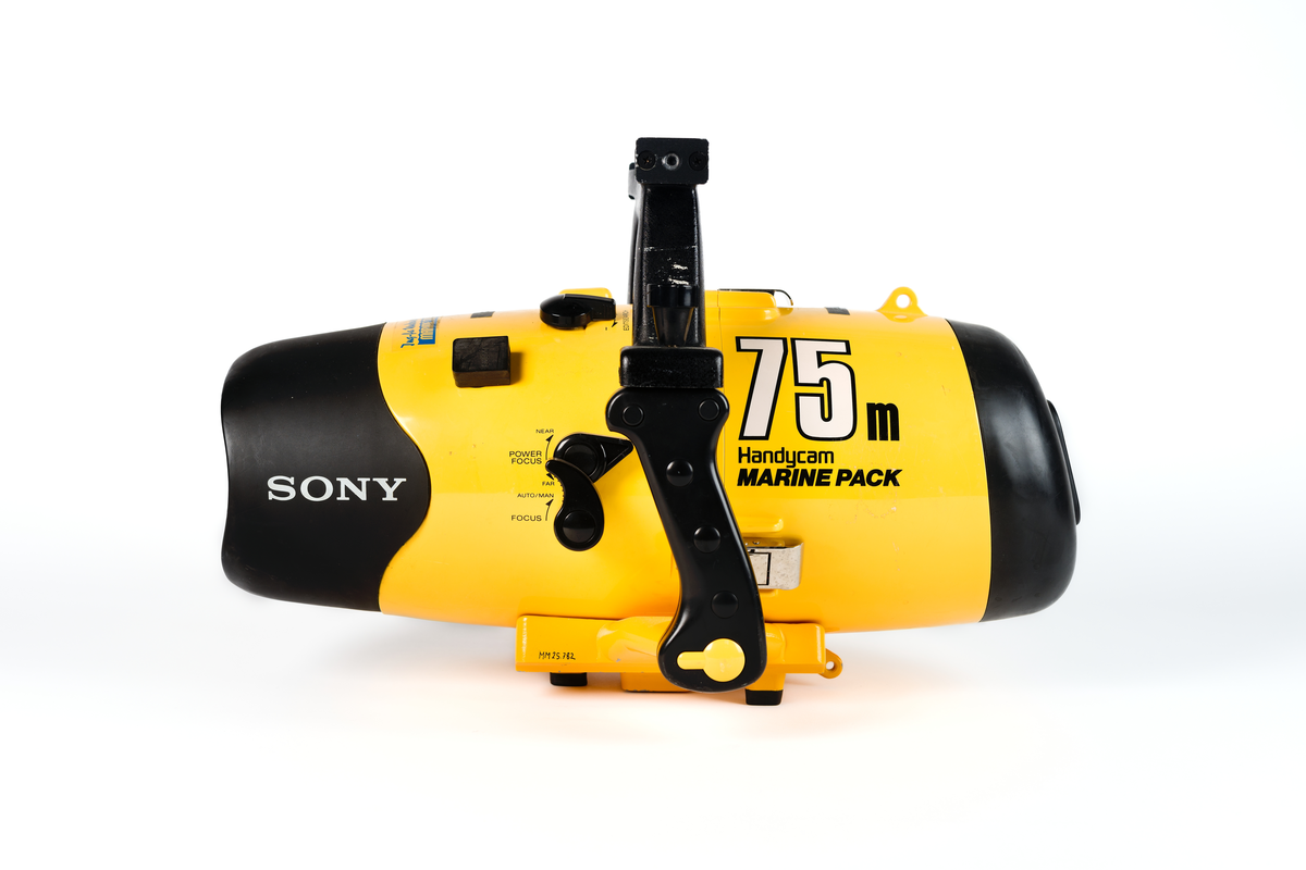 Videokamera i undervattenshus. Gult plasthölje, cylinderformat, med svarta vred och svarta handtag. Rött inspelningsvred samt röd elkontakt. Märkt: "Sony, 75 m Handycam Marine Pack." Dymolapp på höljet med text: "Koster". I höljet en videokamera i svart plast. "Sony Handycam", samt en monitor kopplad till materan med sladd: "Sony LCD Color Monitor XV-M30E". Monitorn synlig genom glaslucka i höljet. Höljet knäpps fast med tre knäpplås.