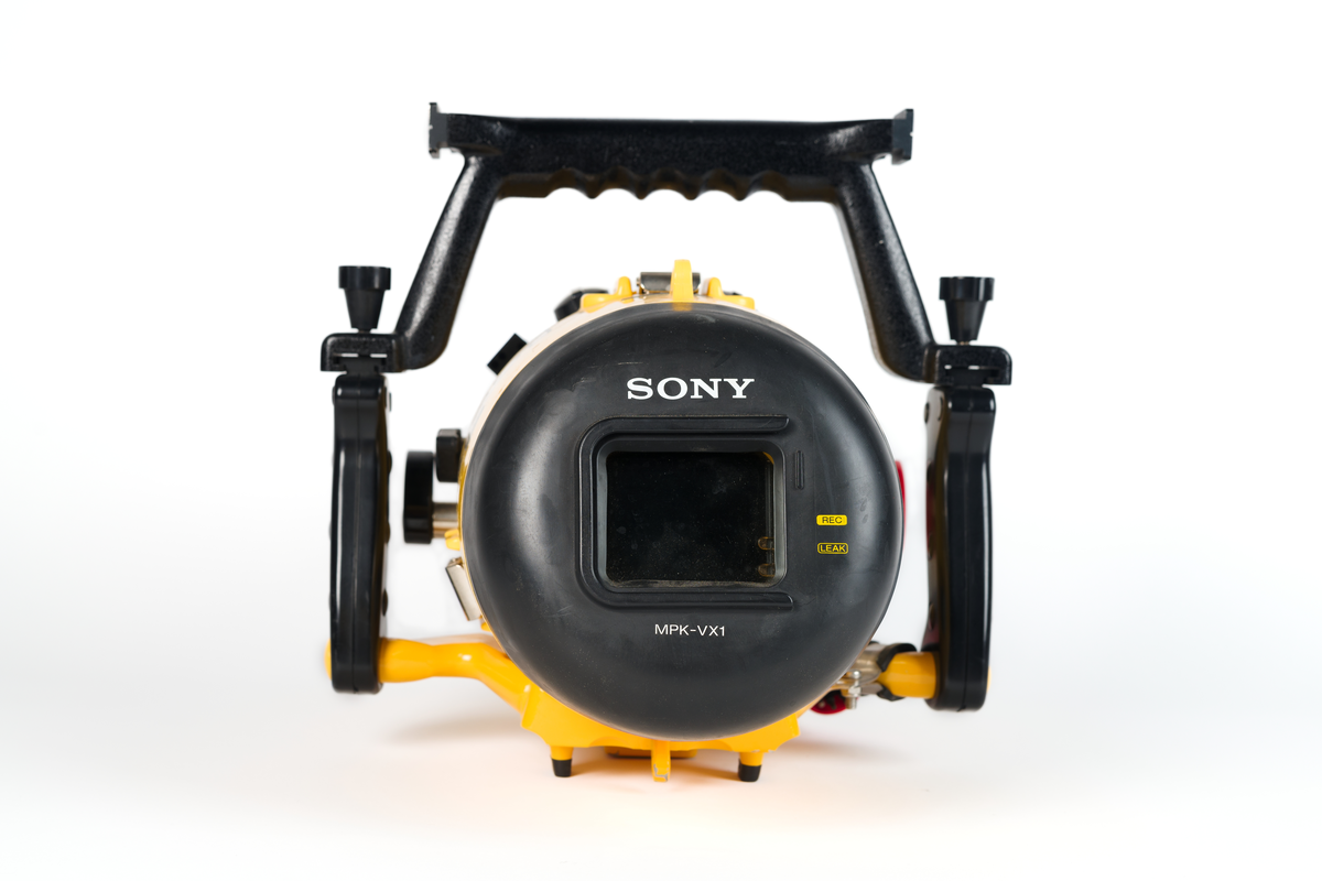 Videokamera i undervattenshus. Gult plasthölje, cylinderformat, med svarta vred och svarta handtag. Rött inspelningsvred samt röd elkontakt. Märkt: "Sony, 75 m Handycam Marine Pack." Dymolapp på höljet med text: "Koster". I höljet en videokamera i svart plast. "Sony Handycam", samt en monitor kopplad till materan med sladd: "Sony LCD Color Monitor XV-M30E". Monitorn synlig genom glaslucka i höljet. Höljet knäpps fast med tre knäpplås.