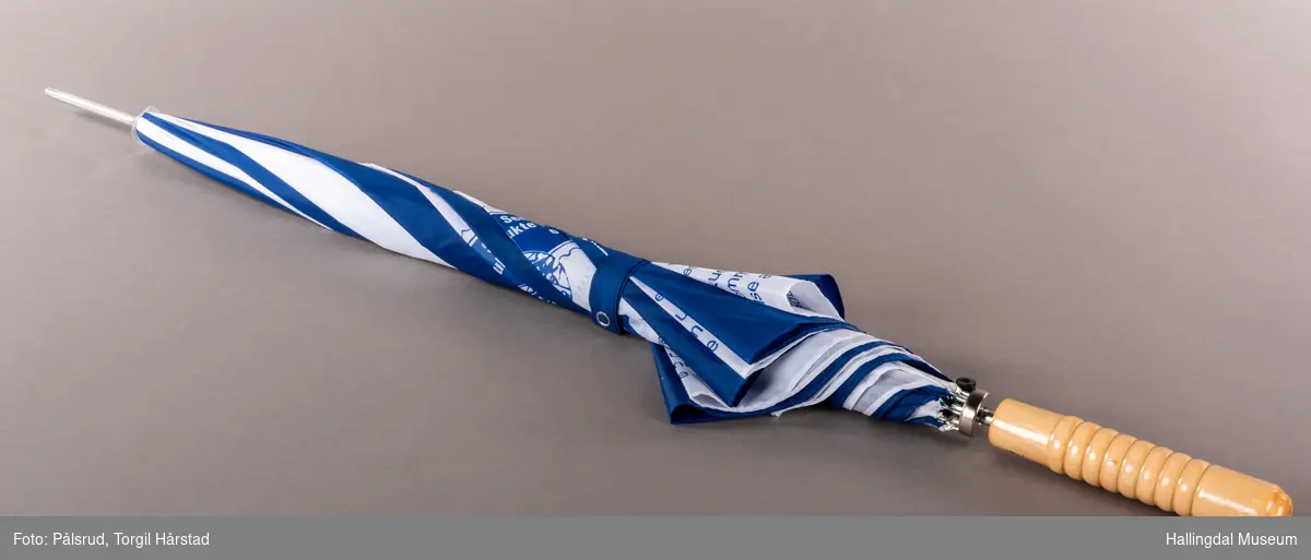 En blå og hvit paraply, annenhver farge på hver stoffdel. De hvite delene har påtrykk med TUBFRIM-logo og tekst. LAng, blank metalltapp i tuppen. Stangen er blank metall med sorte plastikkdeler. Håndtaket er av tre.