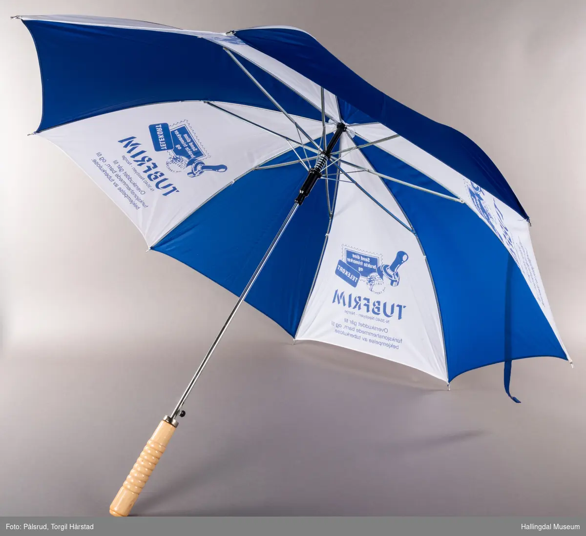 En blå og hvit paraply, annenhver farge på hver stoffdel. De hvite delene har påtrykk med TUBFRIM-logo og tekst. LAng, blank metalltapp i tuppen. Stangen er blank metall med sorte plastikkdeler. Håndtaket er av tre.