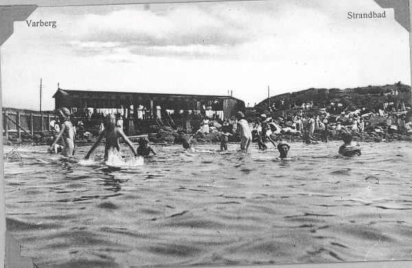 Vykort, "Varberg. Strandbad". Badande med badhytterna i bakgrunden vid Skarpe Nord, då damernas badplats och sedermera nakenbad. Stadens styrande ville få folk att använda badkläder på 1910-talet, så hytternas badföreståndare hyrde ut sådana. På hytthusets räcke hänger badhandukarna på tork och påklädda badbiträden står och ser ut över de badande. Mot 1920-talets slut hade badplatsen utvecklats mer till ett nakenbad för kvinnor.