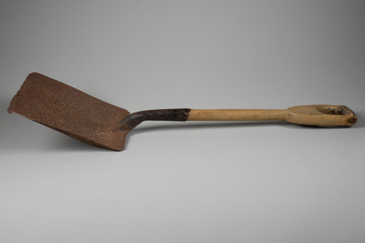 Firkantet fyrbøterspade i jern med malt grønn/brunfarget treskaft .
Jernet er rustent.
Merke øverst på treskaft både på for og bakside