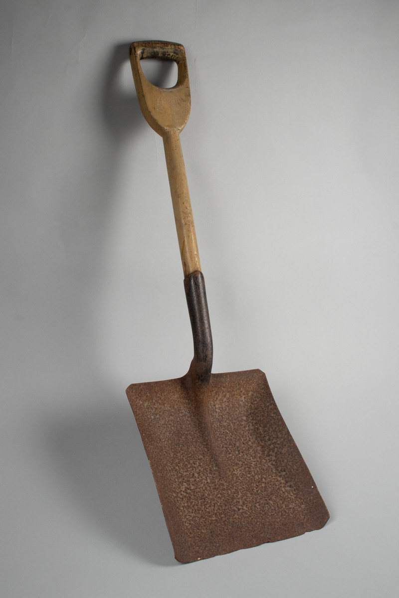 Firkantet fyrbøterspade i jern med malt grønn/brunfarget treskaft .
Jernet er rustent.
Merke øverst på treskaft både på for og bakside