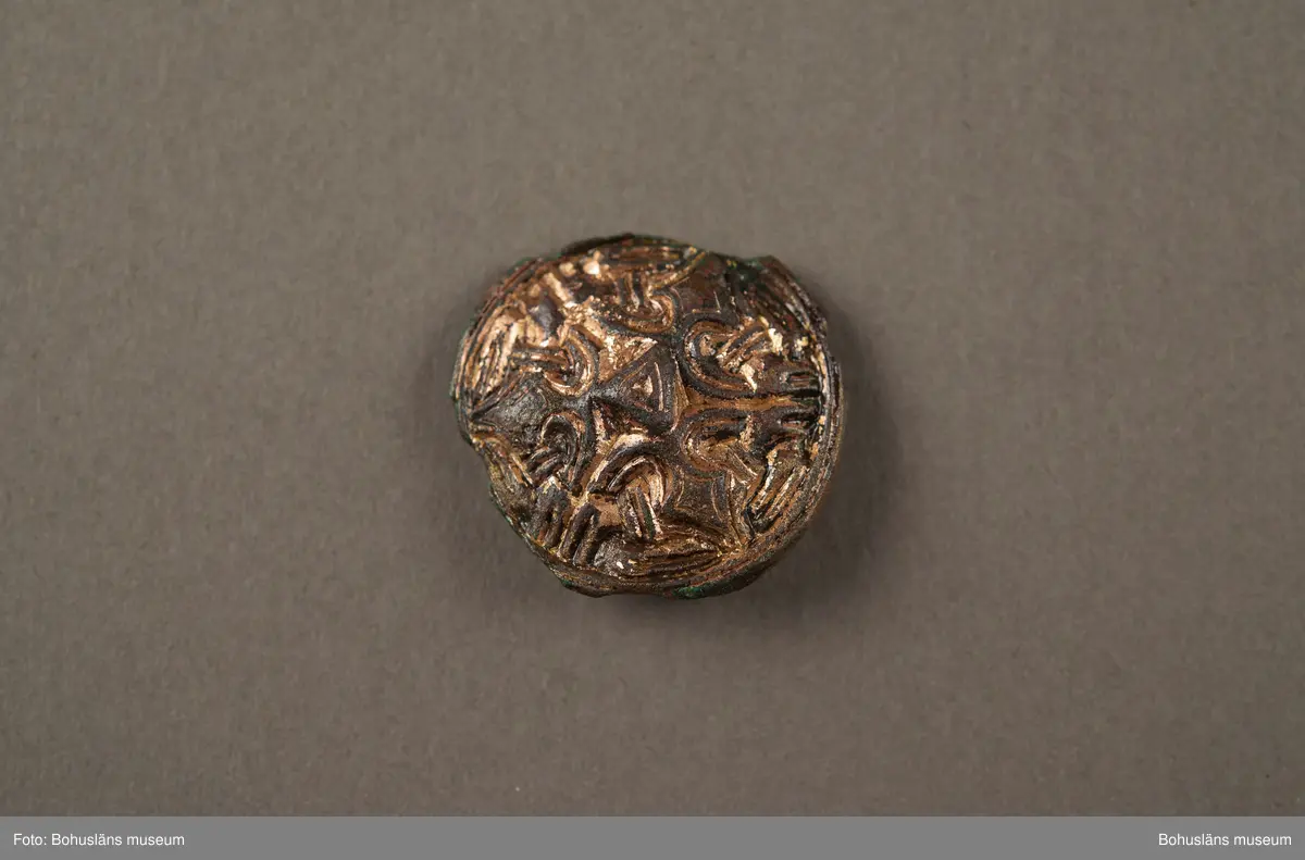 Spänne av brons, runt, med gjuten ornamentik, diameter ca 29 mm. Föremålet är något skadat. Förgyllningen har en rödare ton, vilket tyder på att det är en guldlegering med rätt högt kopparinnehåll. Nålen på baksidan saknas.

Spännet är av en typ som brukar dateras till yngre järnålder, vikingatid. Det hittades år 1878 på gården Trättestad i Kville socken och skänktes till museet år 1879 av soldaten P. A Sörqvist från Söbbskinn i Kville.

Enligt äldre kataloguppgifter i museets arkiv hittades spännet tillsammans med en täljstensgryta (UM002768) och en "gnidsten" (förmodligen avses en slip- eller glättsten).
I fornlämningsregistret för Bohuslän, Kville socken, står att läsa i ett tillägg om fornlämning RAÄ 276 på fastigheten Trättestad: År 1878 erhöll Uddevalla museum från en av högarna härstädes: en täljstensgryta med järngrepe, ett litet runt spänne av brons och en "gnidsten". Enligt uppgift av en åbo i Trättestad hittades i samma hög två "sländtrissor".

Antagligen kommer föremålen från en numera förstörd gravhög inom det gravfält (Kville 276) som idag består av åtta synliga gravar. Kombinationen av föremålen i gravfyndet skvallrar om att det kan ha varit en kvinna som varit begravd i högen. De två sländtrissorna och "gnidstenen" finns idag inte i museets samlingar. Troligen har de aldrig lämnats in.

Ur handskrivna katalogen 1957-1958:
Del av bronshalsprydnad
Diam. 2,9. Bronsspänne m. gjuten ornamentik. Skadat. Trellestad, Kville Bohusl. 1878  4

Lappkatalog: 1:27

Efter okulär besiktning i mikroskåp konstaterades att spännet var förgyllt med rödaktig guld (hög kopparhalt). 2008-11-07/IL

Landskap BOHUSLÄN