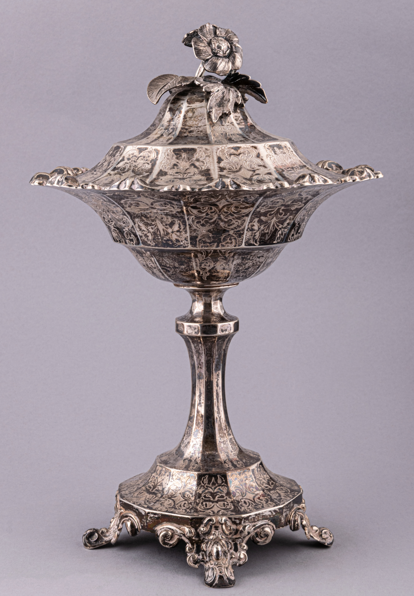 Lock till sockerskål.
Det av ett set om två strösockerskålar i silver, nyrokoko, med skedar. Skål och lock består av tolv pressade delar. Tolvkantig fot med bladformade fötter. Vid skål med kant av pressad bladornamentik. På det paviljongliknande locket sitter en naturalistisk blomknopp samt ett uttag för en sked. Hela sockerskålen utom benet är täckt med graverad bladornamentik. 
Stadstämpel: Stockholm, Mästarstämpel: Gustaf Möllenberg/ Féron.
Årsstämpel: 1869.
Sked, 2st. skaft i Olgamodell. Bladet , snäckliknande och perforerat. Stämplat med föregående.