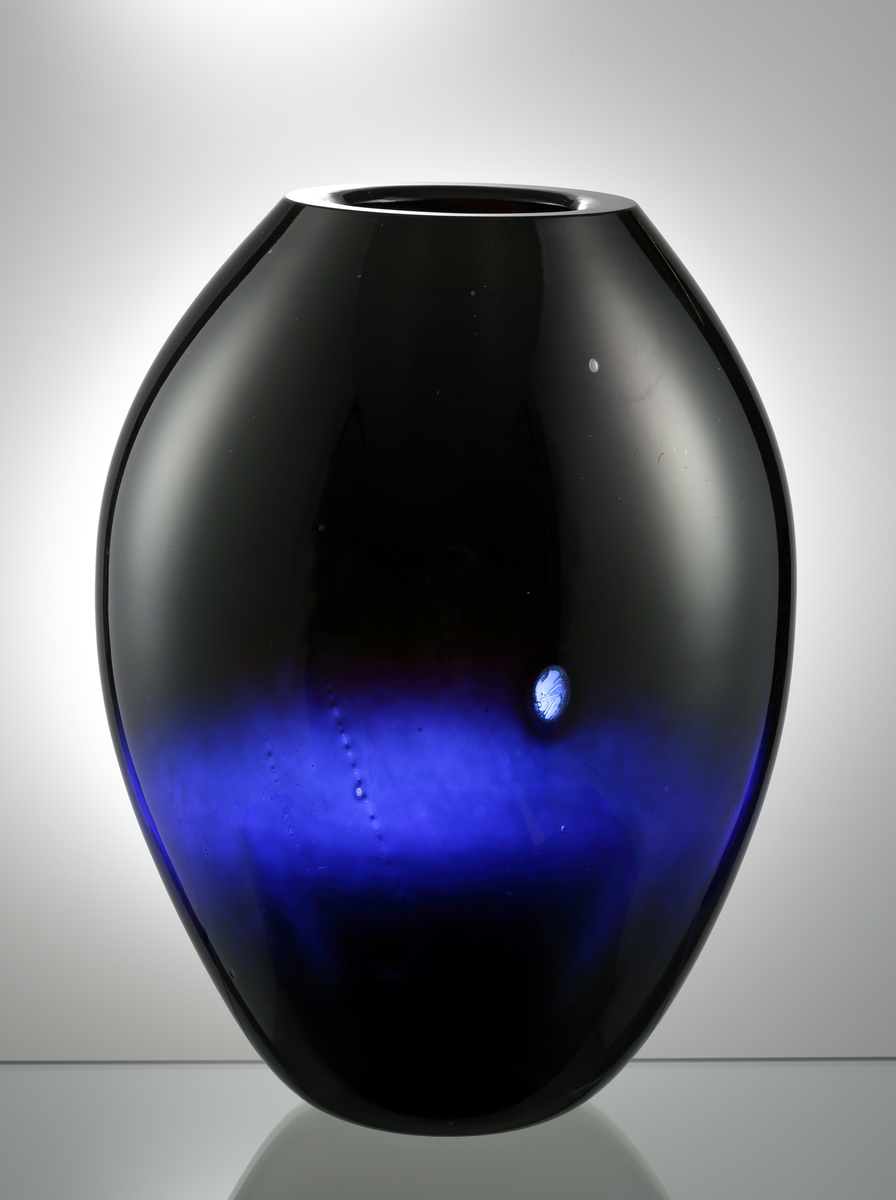 Formgiven av Edwin Öhrström. Stor, tjockväggig vas. Äggformad i mörkblå glasmassa.