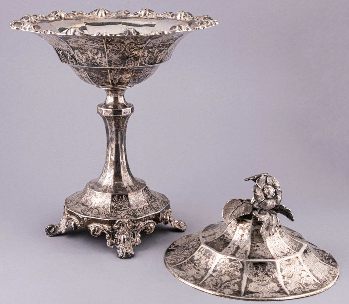 Lock till sockerskål.
Det av ett set om två strösockerskålar i silver, nyrokoko, med skedar. Skål och lock består av tolv pressade delar. Tolvkantig fot med bladformade fötter. Vid skål med kant av pressad bladornamentik. På det paviljongliknande locket sitter en naturalistisk blomknopp samt ett uttag för en sked. Hela sockerskålen utom benet är täckt med graverad bladornamentik. 
Stadstämpel: Stockholm, Mästarstämpel: Gustaf Möllenberg/ Féron.
Årsstämpel: 1869.
Sked, 2st. skaft i Olgamodell. Bladet , snäckliknande och perforerat. Stämplat med föregående.