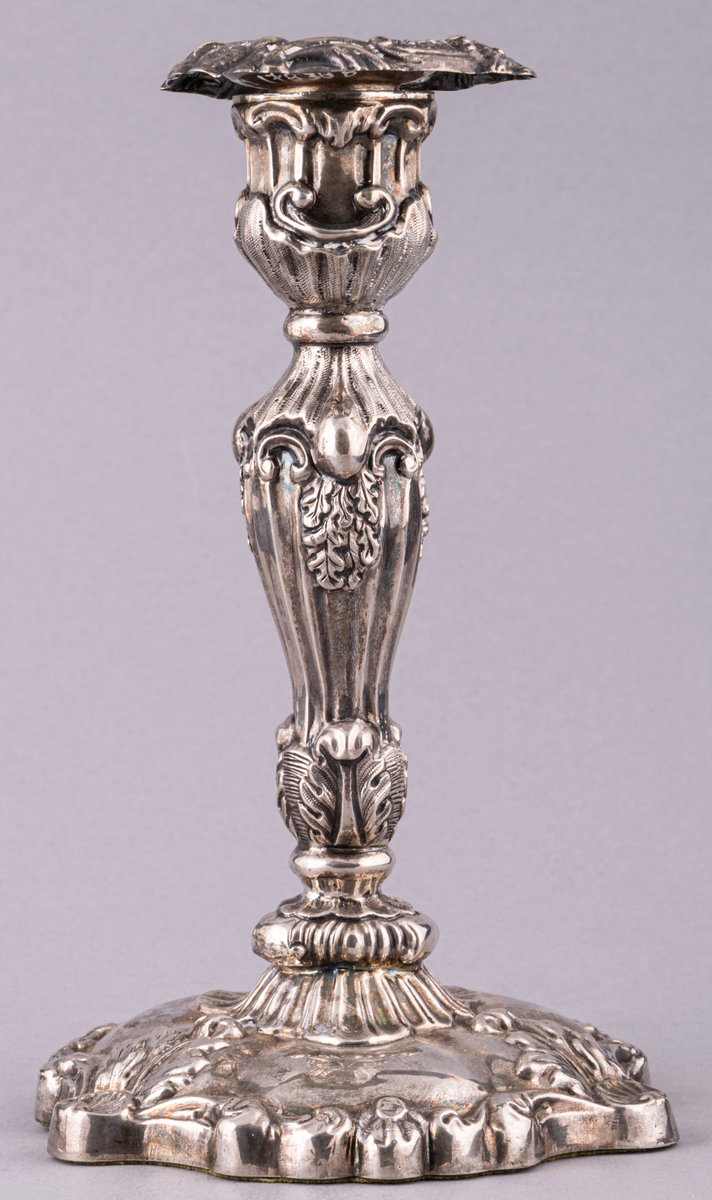 Ljusstake, silver, fotens kant något uddig. Fonden med tre rocailler samt växtornament. Ljuspipan blomprydd.
Ingår i ett par.

Ljuspipa, stämplad med bl.a. kattfot och GM.
Tillverkad Stockholm, Gustaf Möllenborg,
stake 1865, ljuspippa 1863.
Stil: Nyrokoko.