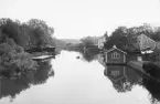 Från Stångebro med kameran riktad söderut fångade fotografen denna vy. Året var 1900 och Stångån bildade då som senare gräns mellan staden Linköping och dess omland. Till vänster i grönskan skymtar roddklubbens båthus och längre bort stadens bägge kallbadhus. Närmast till höger den strandnära bebyggelsen till Linköpings arbets- och fattighus.