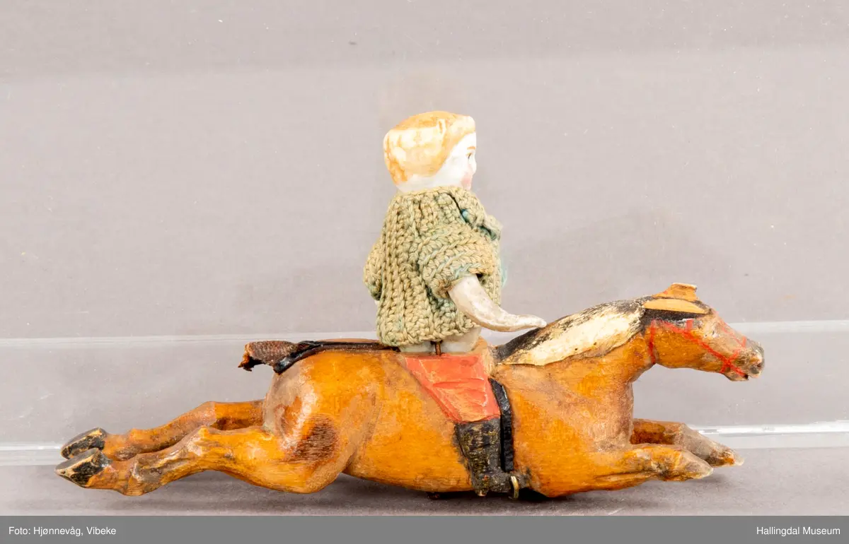 Utskåren hest med påmontert porselendokke med heklet ulljakke. Hesten i full galopp/sprang. Hull midt under tyder på at det har vært montert på et stativ, ant. en karusell med flere figurer enn disse to HFN 09249 a og HFN 09249 b.
