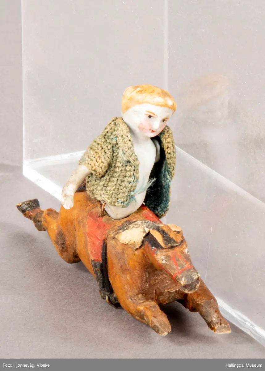 Utskåren hest med påmontert porselendokke med heklet ulljakke. Hesten i full galopp/sprang. Hull midt under tyder på at det har vært montert på et stativ, ant. en karusell med flere figurer enn disse to HFN 09249 a og HFN 09249 b.