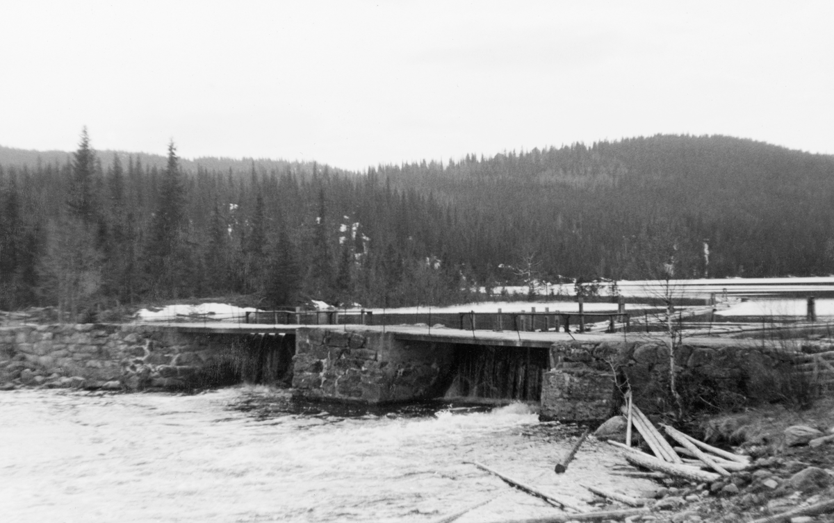 Ringsjødammen i Snertingdalen, fotografert fra nordre elvebredd våren 1938. Ringsjøen er over fire kilometer lang og har et vannspeil som er over en kvadratkilometer stort. Herfra rant Stokkeelva østover mot Mjøsa ved Stokke-gardene. På denne strekningen var elva kommunegrense mellom Biri og Vardal. Til tross for at Stokkeelva bare er om lag halvannen mil lang forteller Gunnar Sætren (1843-1928) i boka «Beskrivelse af Glommen» (1904) at elveløpet var grunt og steinete, slik at det ofte var ganske store tømmermengder som ble «indeliggende» - som altså ikke lot seg fløte ned til Mjøsa i løpet av en sesong. Denne dammen, som ikke nevnes i Sætrens bok, ble sannsynligvis bygd for å magasinere vann i Ringsjøen, som kunne tappes i fløtingssesongen slik at elvefaret ikke ble grunnere enn at tømmeret holdt seg flytende. Dette var en steindam med to løp som begge hadde lukestengsler. Vi vet foreløpig ikke når dammen ble bygd, men i 1916 ble det fremmet krav om at Stokkeelvens fellesfløtningsforening skulle reparere dammen og erstatte skader vannet og tømmeret hadde forårsaket.