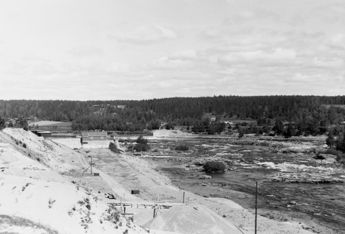 Fra anleggsområdet mellom Skjefstadfoss kraftstaston og damanlegget hvor vannet til turbinene ble hentet fra. Dette var et område med mye sand og grus, der det var bygd en kjøreveg som forbandt de to delene av anlegget. Bak det forholdsvis høye betongbassenget vannet til kraftverket ble hentet fra, mer til høyre i bildet, ser vi overløpsdammen som tømmeret ble fløtet over. I elveløpet nedenfor dammen, til høyre i bildet, ser vi tømmer som har strandet i et grunt parti i skillet mellom østre og vestre elveløp, som deles av Tjuvholmen, som ligger til høyre for høyre bildekant på dette fotografiet.