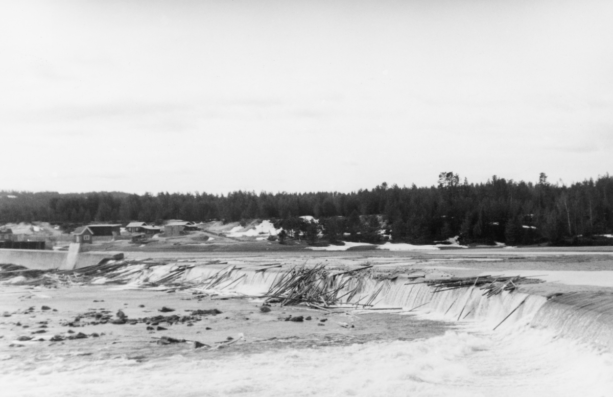 Skjefstadfossen i Heradsbygda i Elverum, fotografert i april 1938.  Fotografiet er tatt fra østsida av elva, mot vest, langsmed den drøyt 300 meter lange oppstuvings- eller løftedammen som magasinerte vann til kraftverket, som lå drøyt 400 meter lengre nede ved vestre bredd.  Lengst til venstre i bildet skimtes kanalmuren ved inntaket til de digre turbinrørene og noen bakenforliggende hus.  Fotografiet viser ellers hvordan en del fløtingstømmer er blitt liggende på kanten av den nevnte oppstuvings- eller løftedammen. 