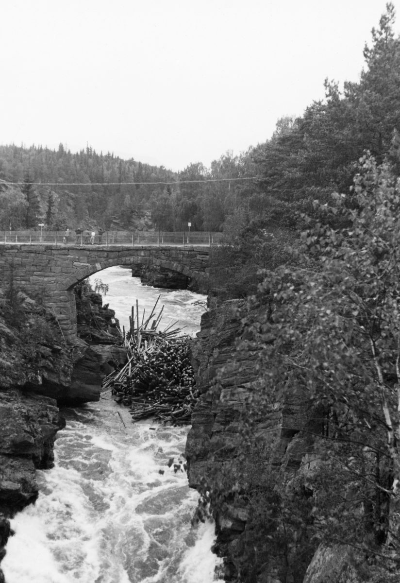 Tømmervase under brua ved Harpefoss i Sør-Fron i Gudbrandsdalen.  Her renner Gudbrandsdalslågen gjennom et trangt gjel, der fløtingstømmeret hadde lett for å sette seg fast.  Da dette fotografiet ble tatt, 10. juni i 1938, lå det en tømmervase like under brua.  Bildet er tatt fra sør mot nord.  Antakelig ble det arbeidet med å løse virket, for vi skimter en del unger som lente seg over brurekkverket og så nedover mot noe som foregikk i elvejuvet.  Harpefossbrua var forbindelsesledd mellom folk som bodde i den lille tettbebyggelsen rundt jernbanestasjonen på østsida av elva og dem som bodde på «Baksida», vest for elva, og dermed litt i skyggen av det forholdsvis bratte terrenget der.  I panikken som bredte seg i aprildagene i 1940 ble brua sprengt ut fra en forestilling om at dette kunne hindre den tyske militære framrykkinga, noe som viste seg å være fullstendig virkningsløst.  Seinere er det bygd ei moderne betongbru på dette stedet.