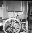 En kvinna sitter vid en spinnrock i ett kök.