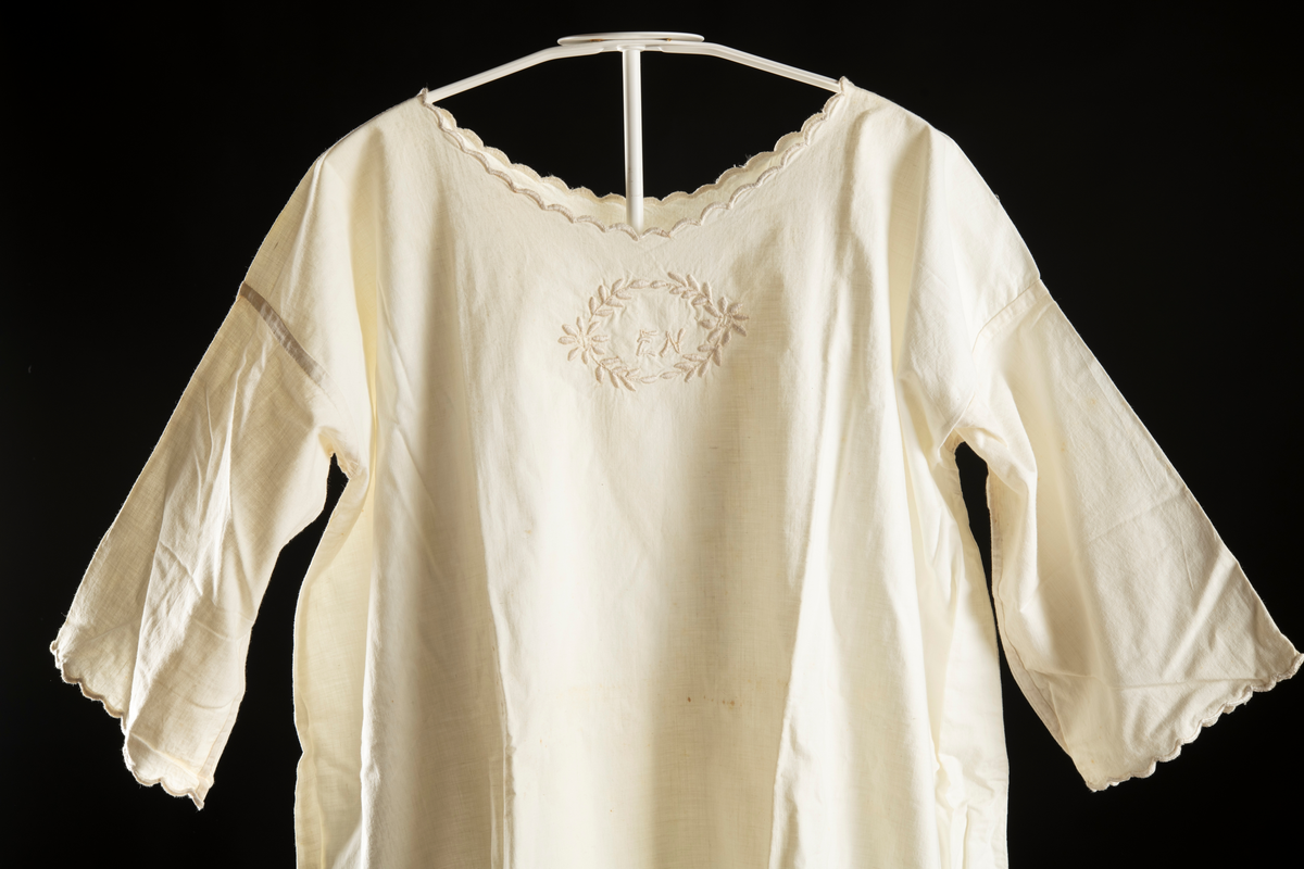 Nattlinne av vitt vävt tyg. Halvlång ärm med broderad kant. Monogram "EN" med krans av blad runt broderat på bröstet. Broderi kring halslinning.