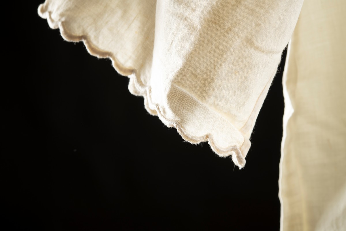 Nattlinne av vitt vävt tyg. Halvlång ärm med broderad kant. Monogram "EN" med krans av blad runt broderat på bröstet. Broderi kring halslinning.