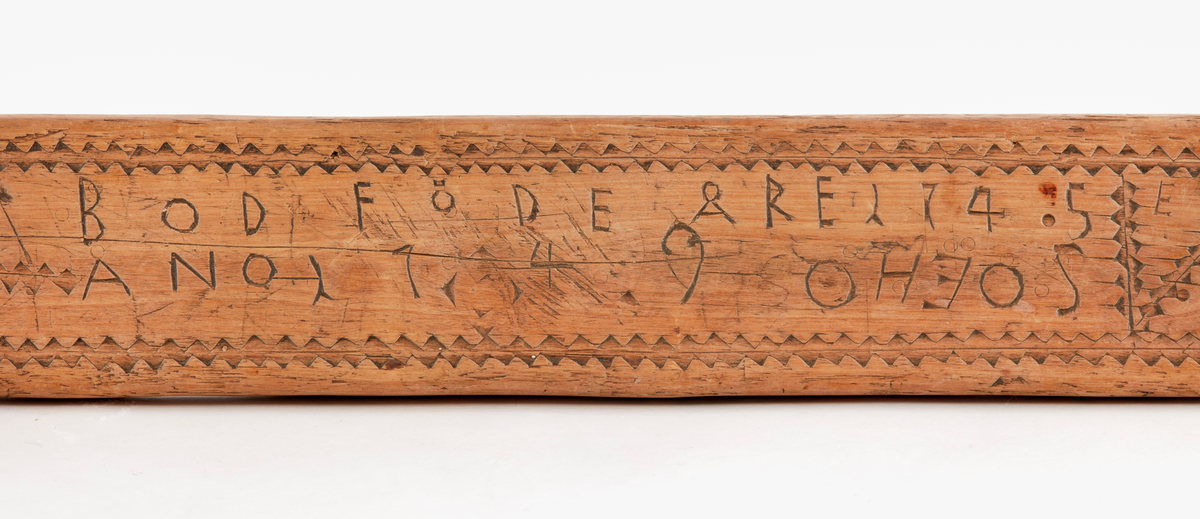 Födelsesticka i form av avlång bräda. Skuren dekor på ovansidan med bl.a. fyra korslagda pilar, dalpilar? Skuren märkning på ovansidan: "BOD FÖDE ÅRE 1745",  "ANO 1749 OHEOS", "E".  Skuren märkning på undersidan "KOD". 
Ev. rester av spikhål i var kortände.