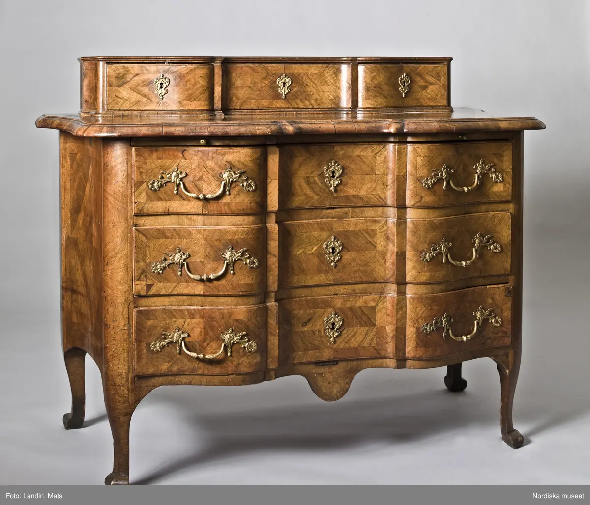 Trelådig byrå, senbarock/Fredrik I:s tid, 1730-40-tal, enkelsvept, med arbalète-formad svängd front. Uppsats med tre lådor. Fanerad på furu med valnöt. Faneret på lådfronterna lagt i kvadrerade fält med fris. Svängda ben med klubbfötter. Utdragbar skrivskiva fanerad med valnöt på ovansidan. Franska régencebeslag av gjuten mässing. Lådbottnar och lådsidor av al. Hela fackbottnar.

Brännstämplad tre gånger på ryggen: "I.H.F" (Johan Hugo Fürloh, mästare i Stockholms snickareämbete 1724-1745. Verkstaden drevs vidare av änkan till 1747).

Anm: Mellersta lådans baksida och botten maskäten. På ryggen etikett med påskrift: "Cyanvätesgasbehandlad  den 26/2 1938 / Hälsovårdsnämndens Cyanvätesanstalter".
Två bygelhandtag nytillverkade juni 2005 på B A konstgjuterier AB, Rosenlundsgatan, Stockholm (märkta med inv.nr och ' Nytillv. 2005' ).  
/Anna Womack 2006-03-02