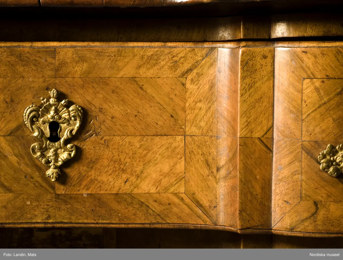 Trelådig byrå, senbarock/Fredrik I:s tid, 1730-40-tal, enkelsvept, med arbalète-formad svängd front. Uppsats med tre lådor. Fanerad på furu med valnöt. Faneret på lådfronterna lagt i kvadrerade fält med fris. Svängda ben med klubbfötter. Utdragbar skrivskiva fanerad med valnöt på ovansidan. Franska régencebeslag av gjuten mässing. Lådbottnar och lådsidor av al. Hela fackbottnar.

Brännstämplad tre gånger på ryggen: "I.H.F" (Johan Hugo Fürloh, mästare i Stockholms snickareämbete 1724-1745. Verkstaden drevs vidare av änkan till 1747).

Anm: Mellersta lådans baksida och botten maskäten. På ryggen etikett med påskrift: "Cyanvätesgasbehandlad  den 26/2 1938 / Hälsovårdsnämndens Cyanvätesanstalter".
Två bygelhandtag nytillverkade juni 2005 på B A konstgjuterier AB, Rosenlundsgatan, Stockholm (märkta med inv.nr och ' Nytillv. 2005' ).  
/Anna Womack 2006-03-02