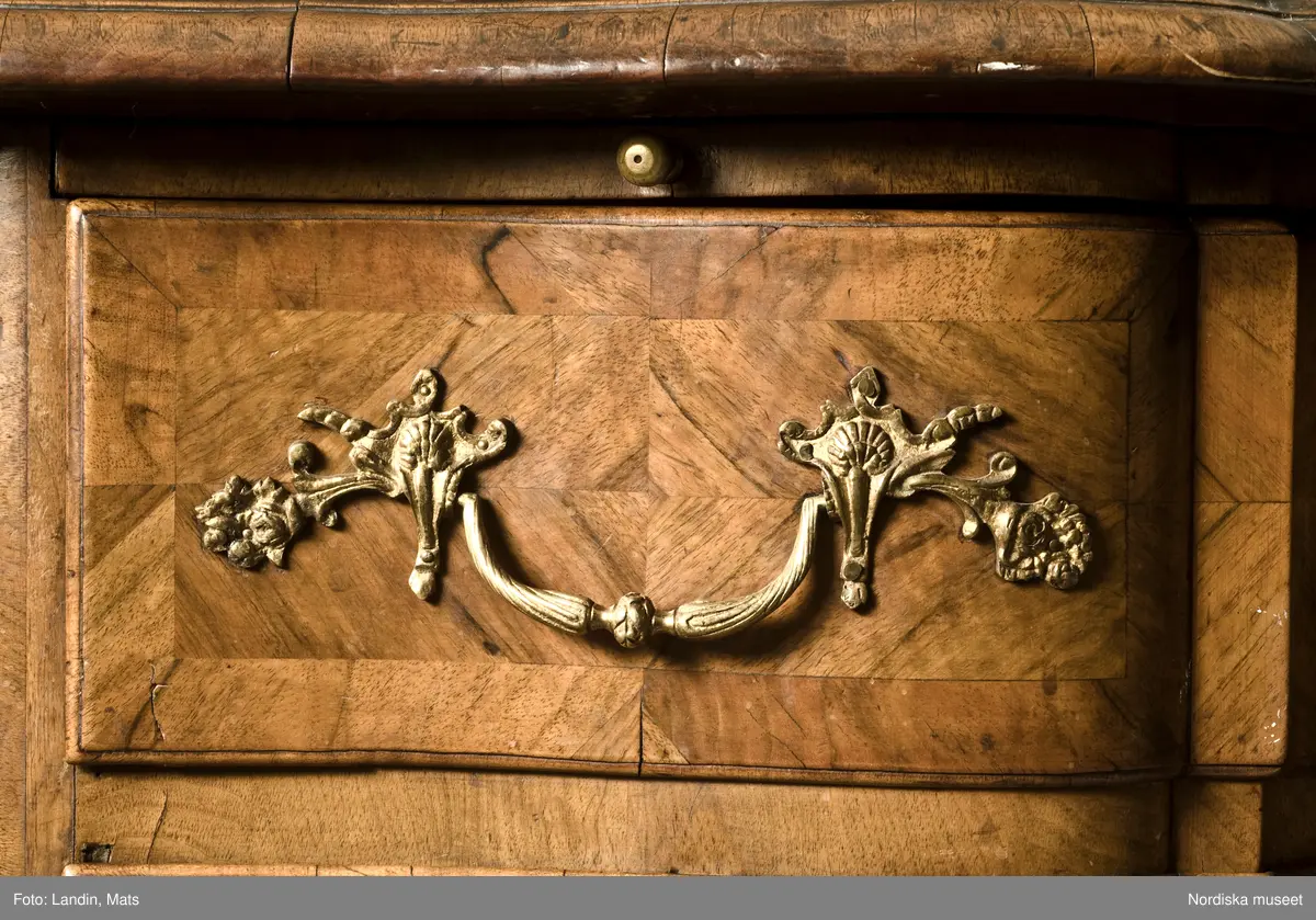 Trelådig byrå, senbarock/Fredrik I:s tid, 1730-40-tal, enkelsvept, med arbalète-formad svängd front. Uppsats med tre lådor. Fanerad på furu med valnöt. Faneret på lådfronterna lagt i kvadrerade fält med fris. Svängda ben med klubbfötter. Utdragbar skrivskiva fanerad med valnöt på ovansidan. Franska régencebeslag av gjuten mässing. Lådbottnar och lådsidor av al. Hela fackbottnar.

Brännstämplad tre gånger på ryggen: "I.H.F" (Johan Hugo Fürloh, mästare i Stockholms snickareämbete 1724-1745. Verkstaden drevs vidare av änkan till 1747).

Anm: Mellersta lådans baksida och botten maskäten. På ryggen etikett med påskrift: "Cyanvätesgasbehandlad  den 26/2 1938 / Hälsovårdsnämndens Cyanvätesanstalter".
Två bygelhandtag nytillverkade juni 2005 på B A konstgjuterier AB, Rosenlundsgatan, Stockholm (märkta med inv.nr och ' Nytillv. 2005' ).  
/Anna Womack 2006-03-02