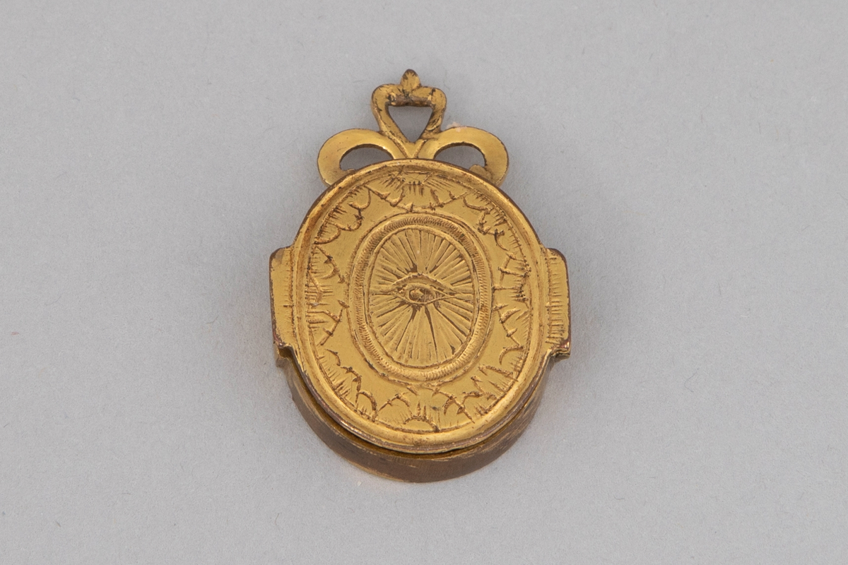 Ovalformet anheng/ medaljong i forgylt metall med hengslet lokk. På innsiden er det et miniatyrmaleri/ trykk av en prest bak glass. Gravert symmetrisk dekor på lokket (øye) og medaljongens bakside (blomst). Øverst på smykket er det tre løkker.