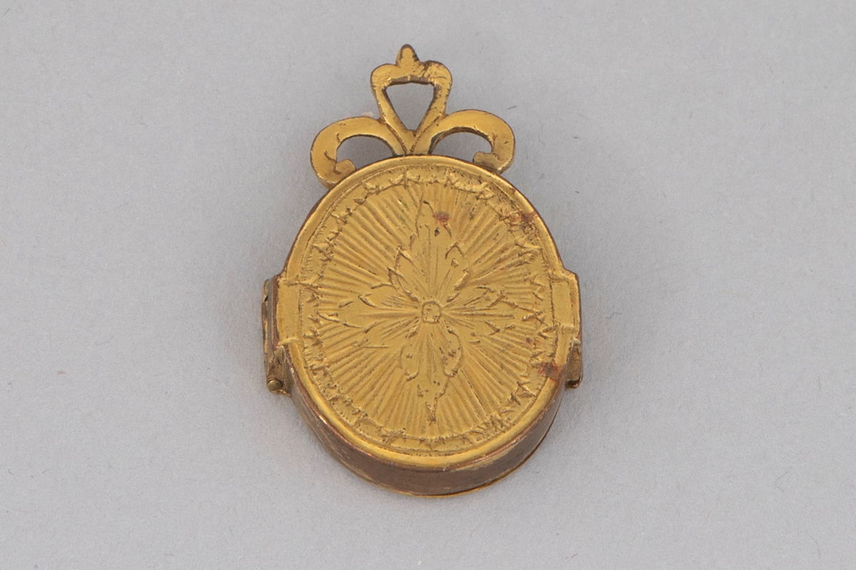 Ovalformet anheng/ medaljong i forgylt metall med hengslet lokk. På innsiden er det et miniatyrmaleri/ trykk av en prest bak glass. Gravert symmetrisk dekor på lokket (øye) og medaljongens bakside (blomst). Øverst på smykket er det tre løkker.