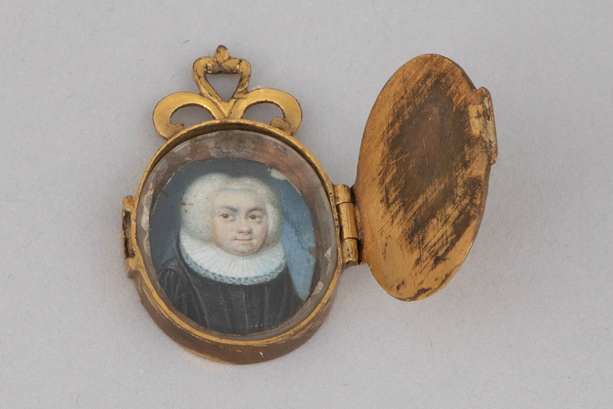 Ovalformet anheng/ medaljong i forgylt metall med hengslet lokk. På innsiden er det et miniatyrmaleri/ trykk av en prest bak glass. Gravert symmetrisk dekor på lokket (øye) og medaljongens bakside (blomst). Øverst på smykket er det tre løkker.