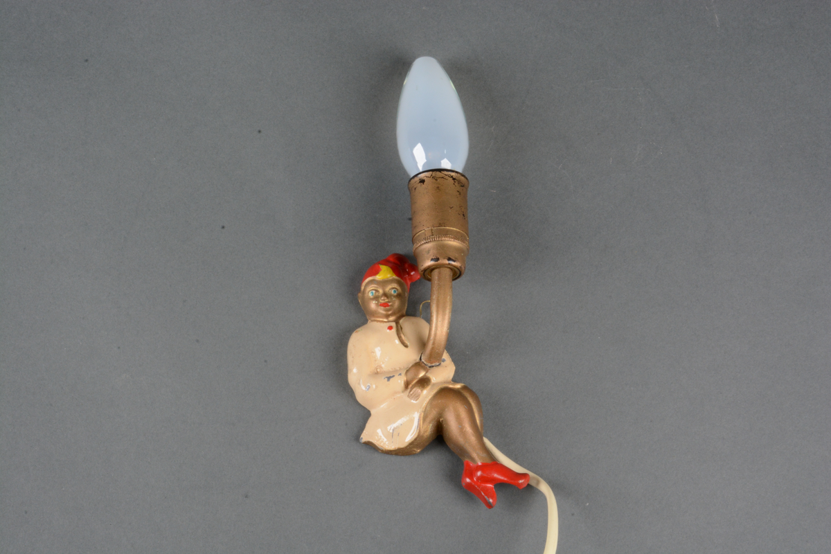 Lampe til å montere på vegg. Lampen (A) er montert på en metallbase som er formet som Ole Lukkøye kledd i nattkjortel, tøfler og nattlue. Ledning uten støpsel. Tilhørende lampeskjerm (B) med kunstfiber og med metallfeste bak. Lampeskjermen er formet som en paraply. Possement rundt kantene på paraplyen.