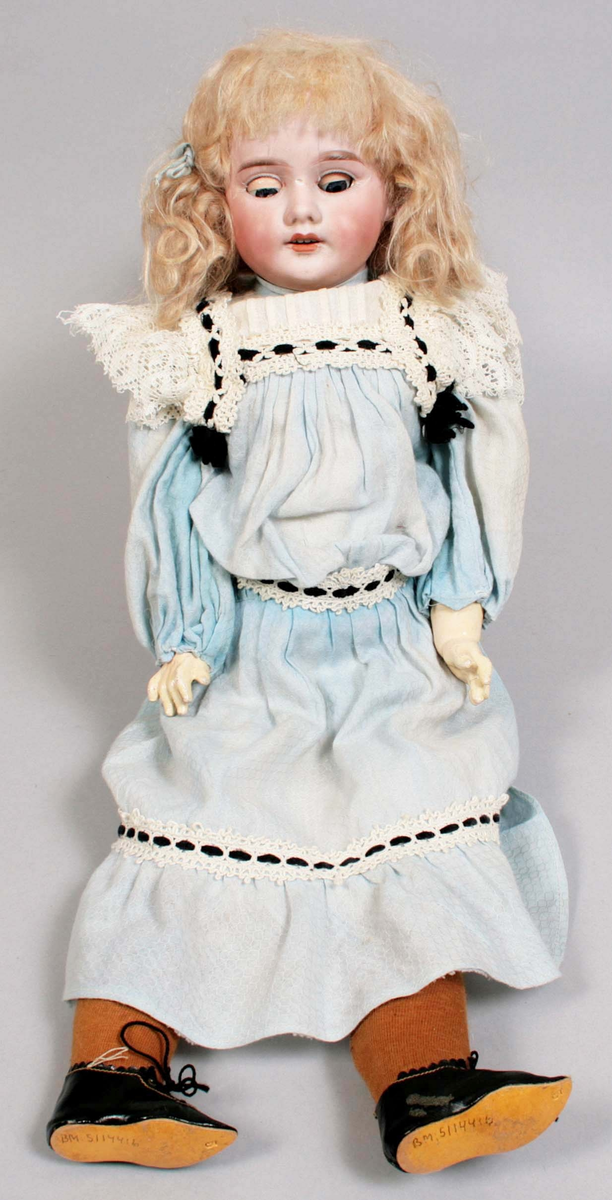 Blunddocka, kallad "Mary". Kropp och  ben av målat trä. Över och underarmar av målad papier maché. Ledade lemmar. Huvud av målat biskviporslin. Rörliga, blå ögon av glas. Blont hår i "page frisyr". Klädd i ljusblå bomullsklänning, dekorerad med vit spets och genomträdda svarta sammetsband. Vit underkjol samt byxor kantade med spets. Bruna knästrumpor i trikå. Svarta skor i konstmaterial. Märkt: "1894 AMDEP made in germany 8".

Tillverkad år 1894, av porslinsmanufakturen "Armand Marseille 1885-1950" i Köppelsdorf, Tyskland.