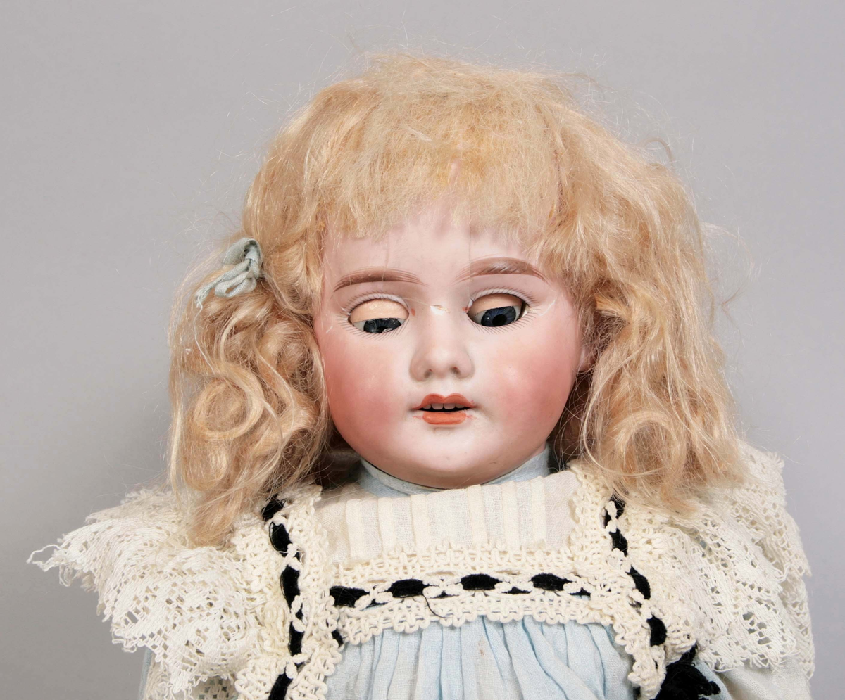 Blunddocka, kallad "Mary". Kropp och  ben av målat trä. Över och underarmar av målad papier maché. Ledade lemmar. Huvud av målat biskviporslin. Rörliga, blå ögon av glas. Blont hår i "page frisyr". Klädd i ljusblå bomullsklänning, dekorerad med vit spets och genomträdda svarta sammetsband. Vit underkjol samt byxor kantade med spets. Bruna knästrumpor i trikå. Svarta skor i konstmaterial. Märkt: "1894 AMDEP made in germany 8".

Tillverkad år 1894, av porslinsmanufakturen "Armand Marseille 1885-1950" i Köppelsdorf, Tyskland.