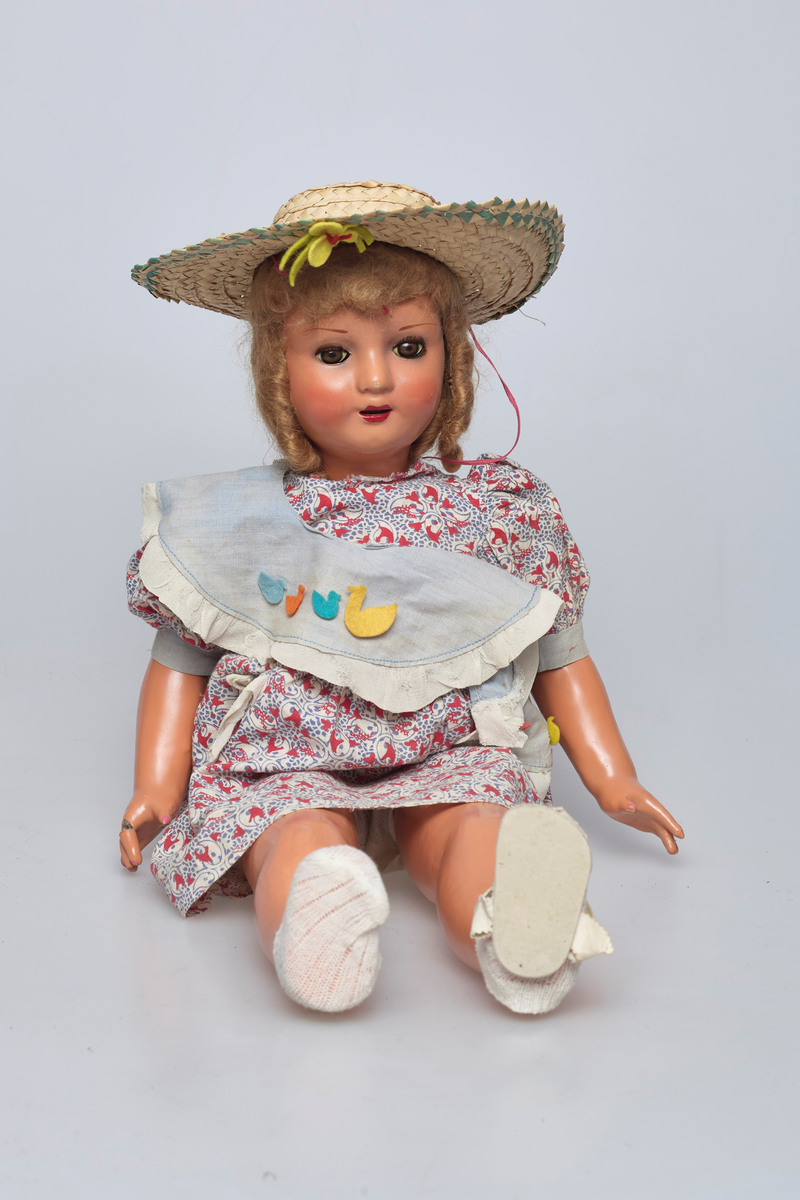 Jentedukke med klær. Kjole og solhatt. Den har en sandal som sitter løst. På kragen er det fire ender av fillt i forskjellige farger.