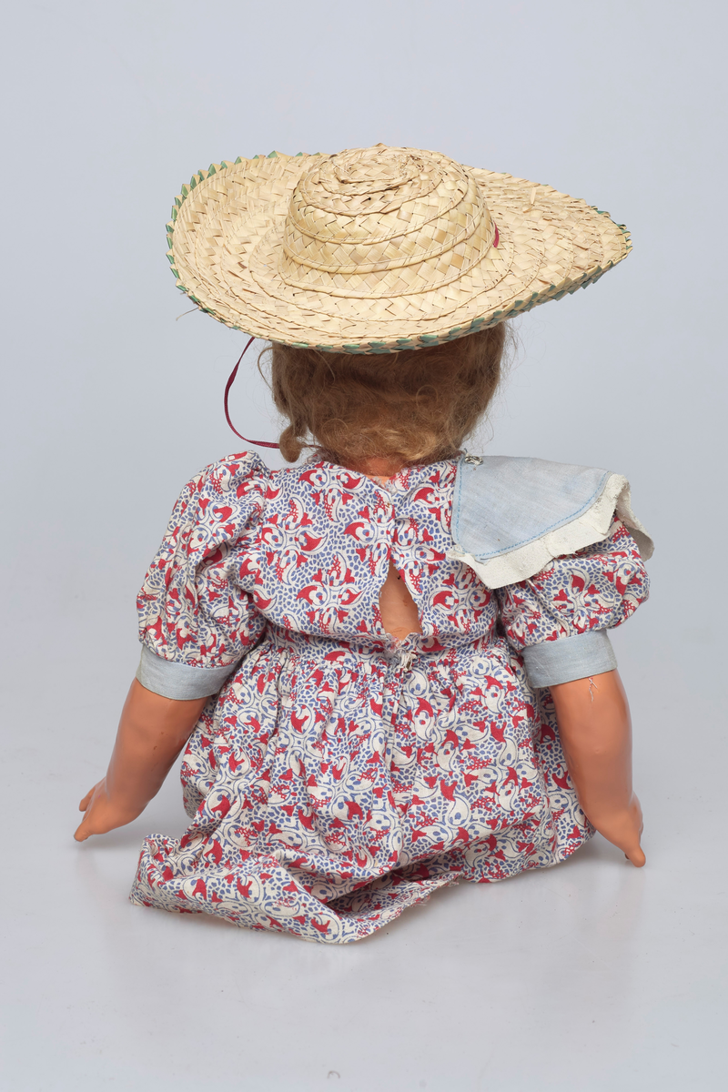 Jentedukke med klær. Kjole og solhatt. Den har en sandal som sitter løst. På kragen er det fire ender av fillt i forskjellige farger.