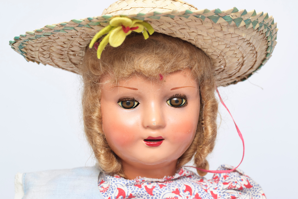 Jentedukke med klær. Kjole og solhatt. Den har en sandal som sitter løst. På kragen er det fire ender av fillt i forskjellige farger.