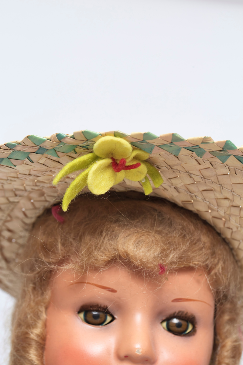 Jentedukke med klær. Kjole og solhatt. Den har en sandal som sitter løst. På kragen er det fire ender av fillt i forskjellige farger.