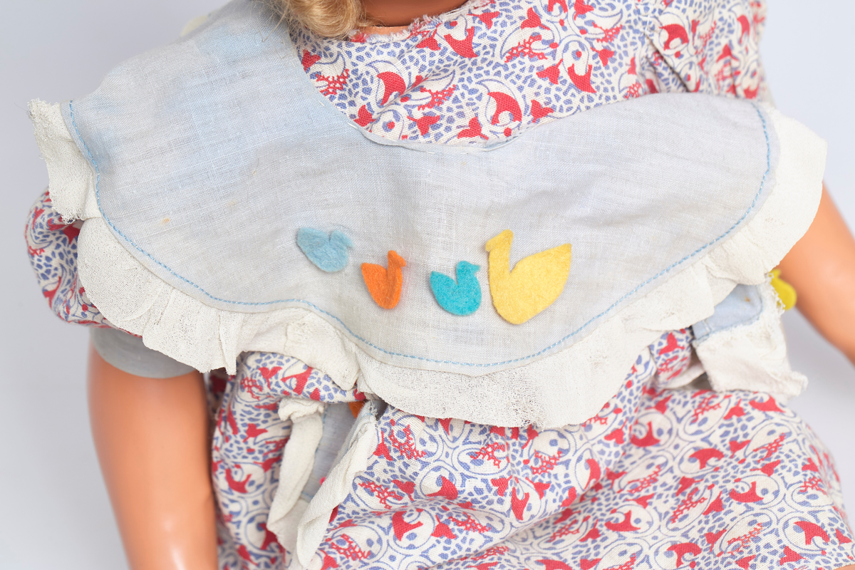 Jentedukke med klær. Kjole og solhatt. Den har en sandal som sitter løst. På kragen er det fire ender av fillt i forskjellige farger.