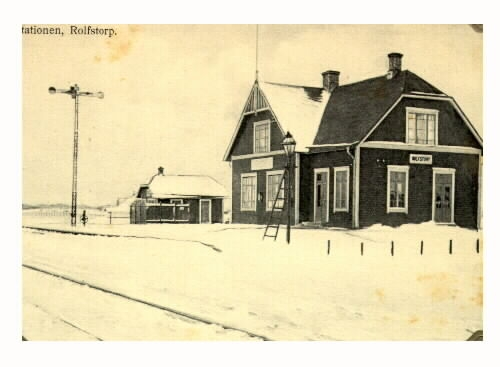 Vykort, "Stationen, Rolfstorp". Vinterbild med järnvägsrälsen i förgrunden, semafor till vänster och en stege rest mot gatlyktan till höger. Stationen ligger utmed Varberg-Ätrans järnväg, som fanns mellan 1911-1961.