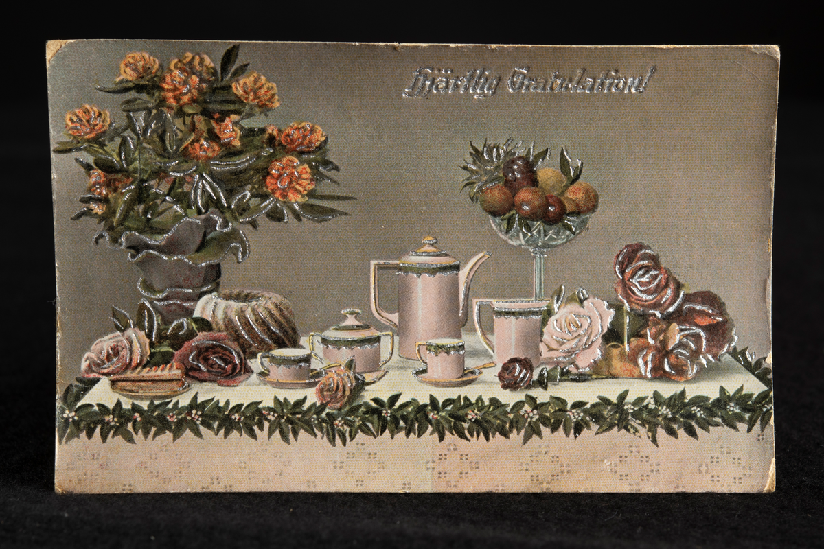 Ett dukat bord, rikt utsmyckat med bladgirland längs kanten av en mönstrad vit duk. Stor blombukett med orange- och rödtonade rosor i gråfärgat papper i vänstra delen, nedanför en bakad, mjuk kaka med vågmönster, samt strödda rosor. I mitten en kaffeservis i vitt porslin, med grön bård. Till höger på bordet ett fruktfat på fot i glas, med blandade frukter, och nedanför en liggande bukett i samma färger som tidigare.
I övre kanten står texten Hjärtlig Gratulation! i silver. Bakgrunden är svagt gråtonad.