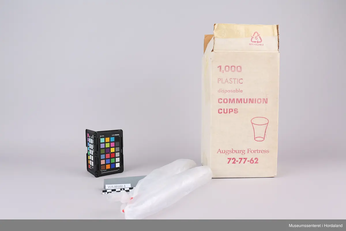 Høy, rektangulær pappeske farget hvit med rød påskrift og rester av tape. Den inneholder femten lange plastposer med femti plastkopper i hver. De har to luftehull og er lukket med rød tape i enden. I tillegg er det en åpnet og knytt rull med nitten plastkopper.