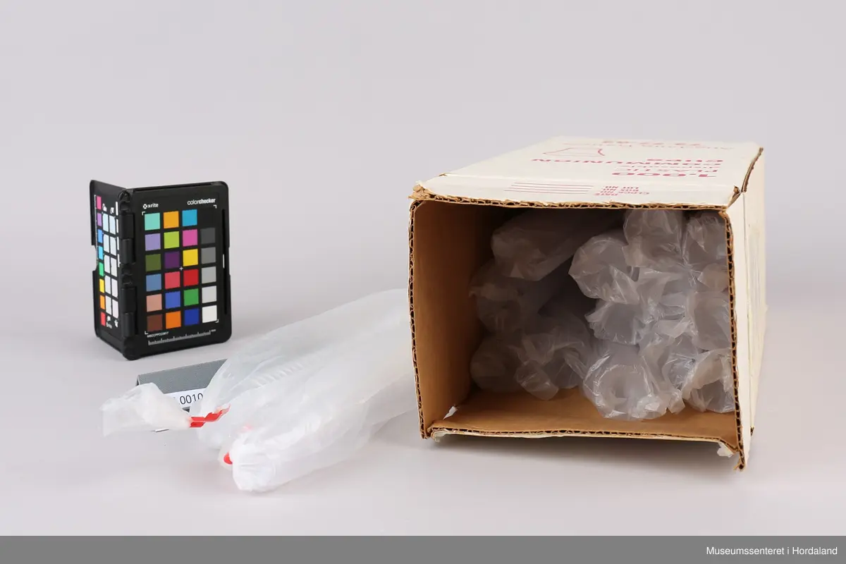 Høy, rektangulær pappeske farget hvit med rød påskrift og rester av tape. Den inneholder femten lange plastposer med femti plastkopper i hver. De har to luftehull og er lukket med rød tape i enden. I tillegg er det en åpnet og knytt rull med nitten plastkopper.