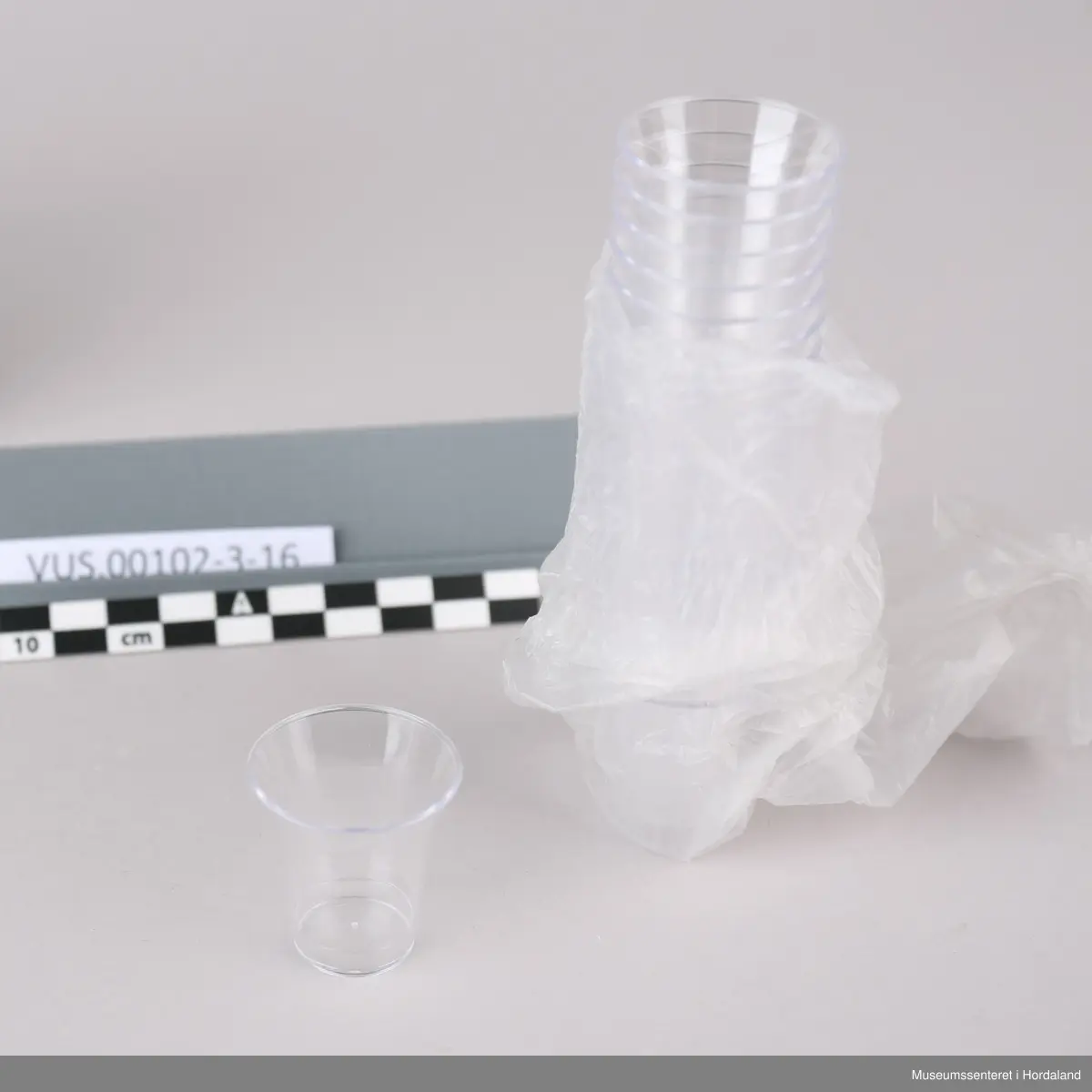 Høy, rektangulær pappeske farget hvit med rød påskrift og rester av tape. Den inneholder femten lange plastposer med femti plastkopper i hver. De har to luftehull og er lukket med rød tape i enden. I tillegg er det en åpnet og knytt rull med nitten plastkopper.