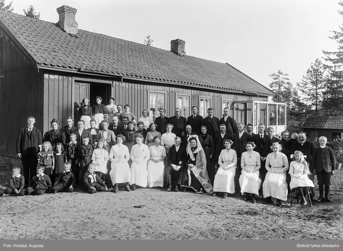 Brudepar og bryllupsgjester fotografert foran forsamlingshus(?). Ukjent sted og ukjente personer. Sort brudekjole. Samme brudepar på ØFB.AKO.00050.