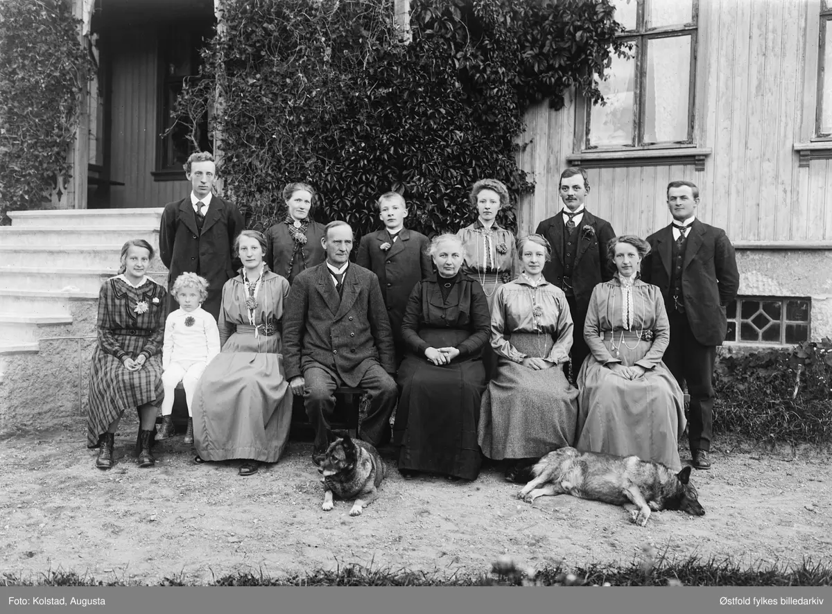 Gruppebilde med familie og to hunder foran inngangsparti, ukjente personer, ukjent sted.