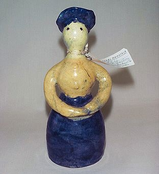 Figur, föreställande en kvinna med blå kjol och hatt. Gul blus och ljusare gult ansikte med bruna ögon.