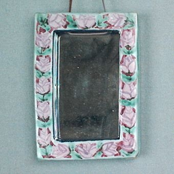 Rektangulär spegel med ram av vitlera, glaserad och målad med ljusgrön färg, rosa rosor och blå kant invändigt samt försedd med läderrem för upphängning.