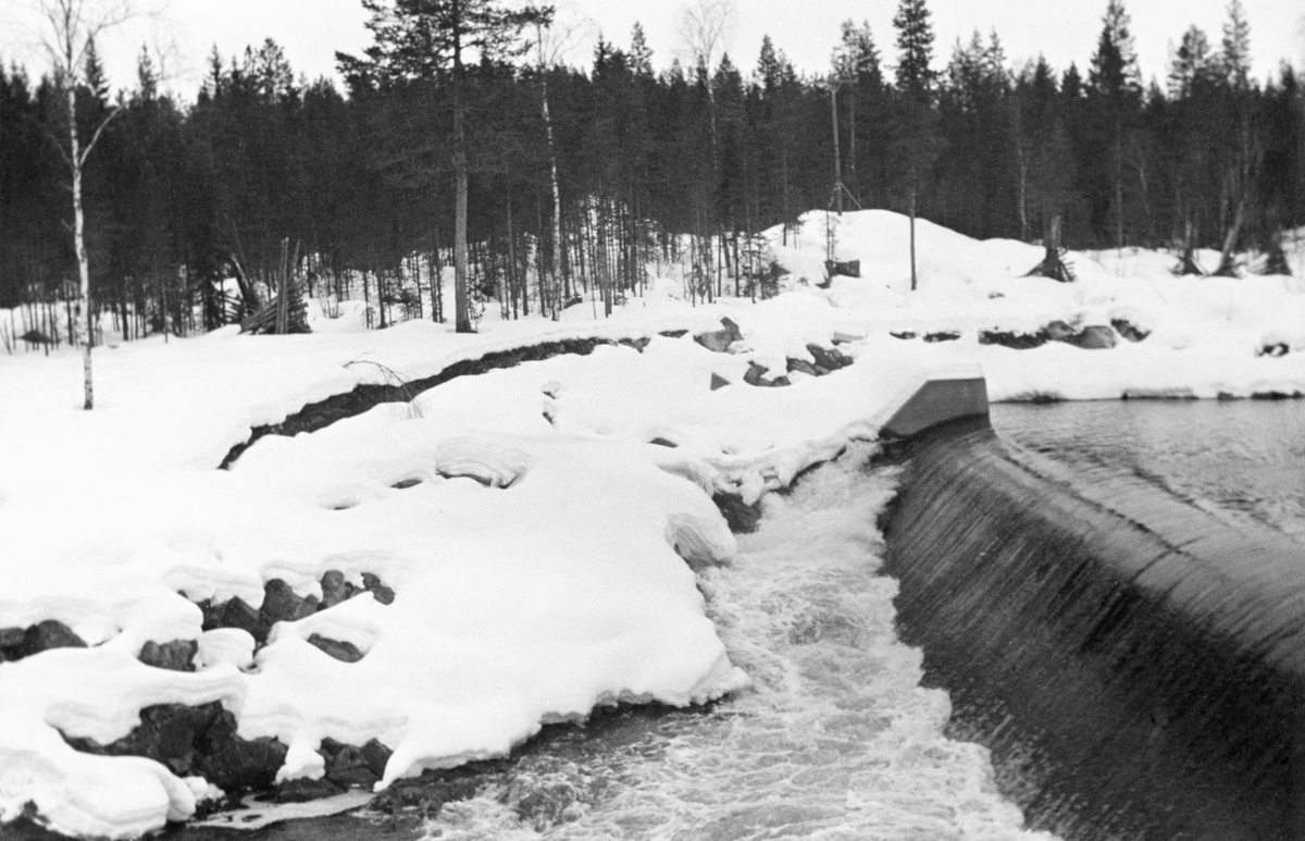 Kraftverksdammen ved Kvernfallet i elva Søndre Osa i Åmot kommune i Hedmark, fotografert i mars 1939, på et tidspunkt da det fortsatt lå snø på marka, men elveløpet var isfritt. Kverfalldammen var en armert betongdam med et avrundet overløp som vannet og - etter hvert - fløltingsviket skulle flyrtet over. Dette bildet ble imidlertid tatt lenge før fløtingssesongen startet. Dammen var bygd mot ei skrå bergflate som tvang vannstrømmen fra nedfallssona sørover før elveløpet igjen dreier mot vest.  På elvebredden til høyre i bakgrunnen ser vi et par plogformete vedreis.