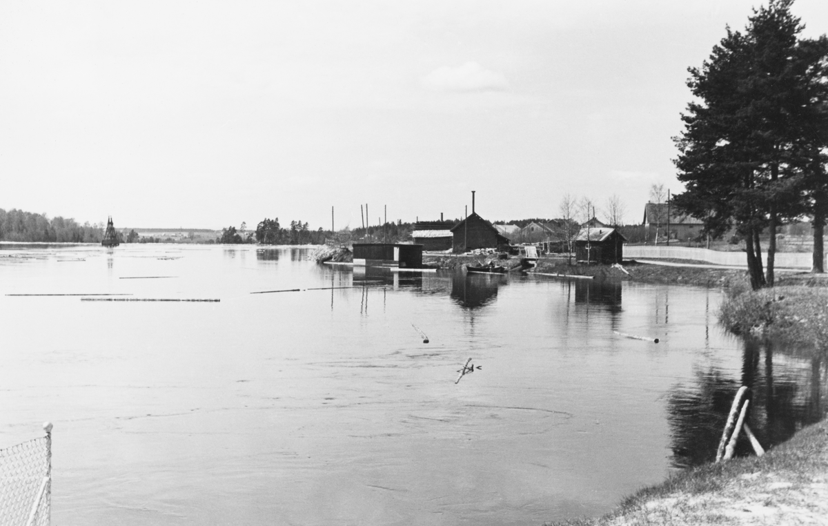 Fra Nabben på Glommas nordre elvebredd like ovenfor Funnefossen i Glomma, i den nordre delen av Nes kommune på Romerike. Fotografiet ble tatt våren 1939, med kameraet vendt i medstrøms retning. Til venstre i bildet, der det blanke, stilleflytende vannspeilet gikk over i et mer opprørt, strømmende fosseløp, sto det et tårn for bærelina på en taubane som ble brukt til å transportere gods fra sørsida av elva, hvor jernbanetraseen gikk, og over til fabrikken på nordsida. Til høyre i bakgrunnen ser vi en del små bygninger, utført i bordkledd bindingsverk, men bak disse skimter vi noen av teglsteinsgavlene i fabrikkomplekset ved Funnefoss Tresliperi.