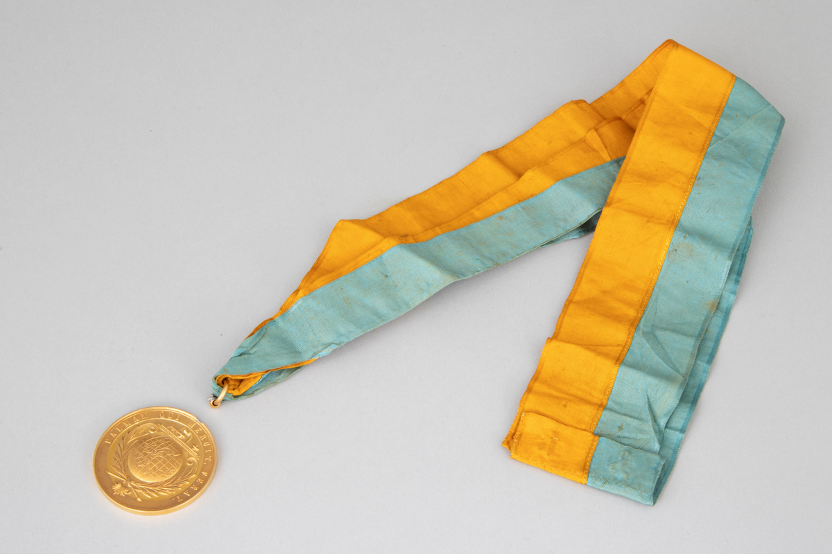 Rund medalje i forgylt metall med opphøyet rand og punslet dekor. På avers er det en globus med palmegrener rundt og en skøyte over. Innskrift langs randen. På revers er det en laurbærkrans, dannet av to kryssende grener med sløyfe rundt, ellers glatt overflate. Langt gult og turkis ripsbånd festet til en ring som er brukket av medaljens kant.
